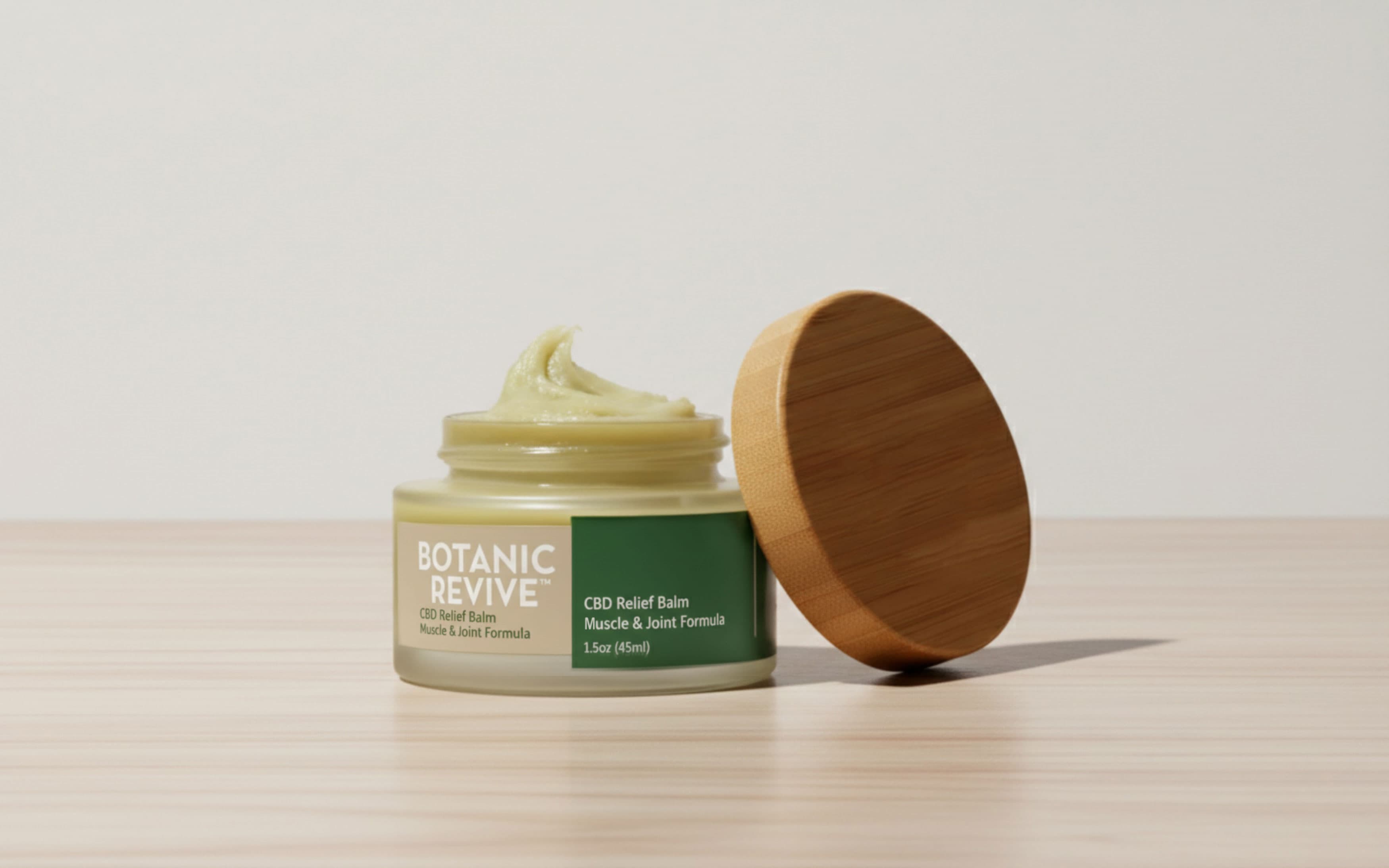 An open glass jar of Botanic Revive CBD Relief Balm showing a thick, pale green ointment swirled at the top. The label indicates a Muscle & Joint Formula in a 1.5oz size, featuring a tan and dark green design. A round bamboo lid leans against the side of the jar on a light wooden surface.