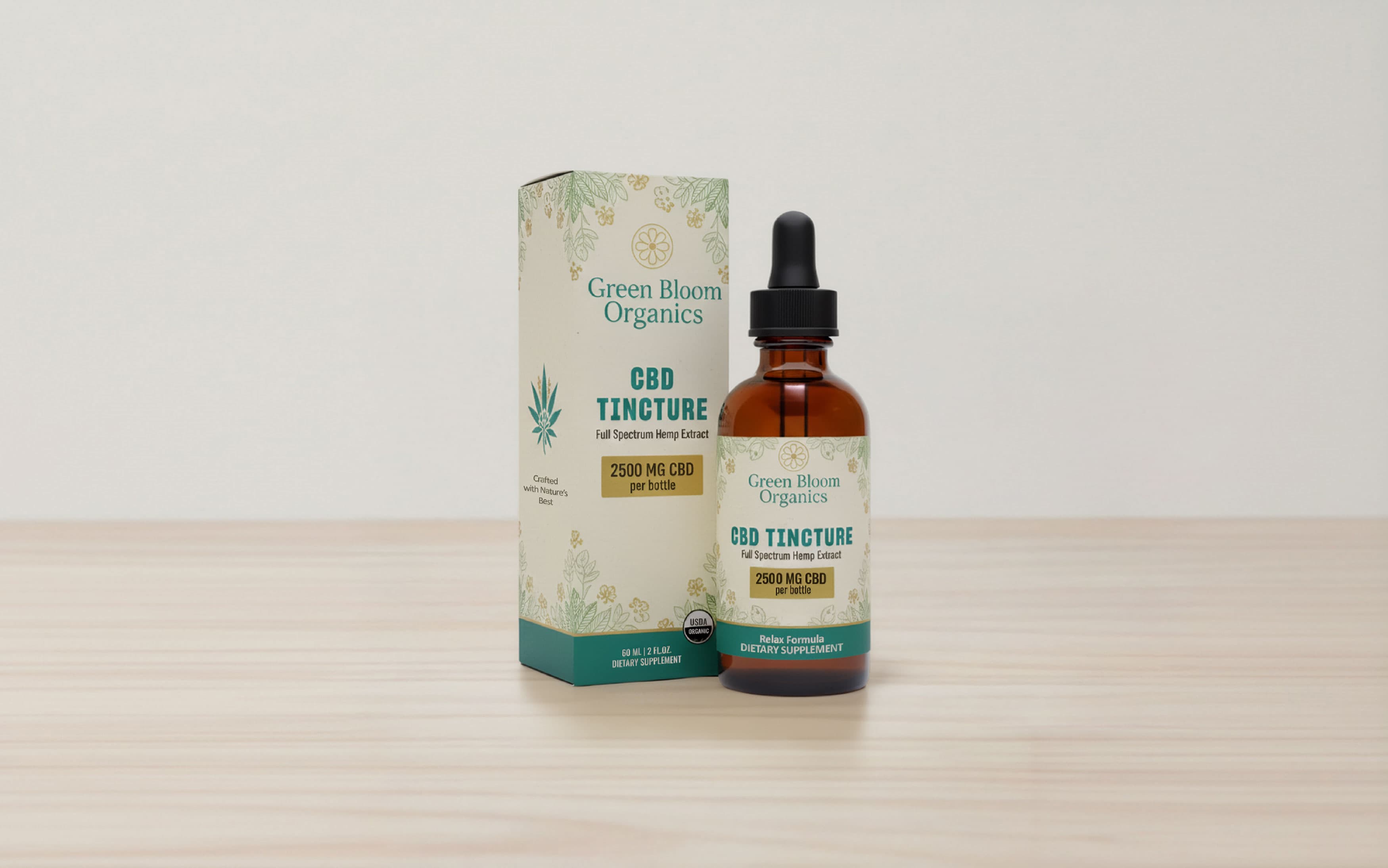 A 60 ml amber glass dropper bottle and its matching cardboard box for Green Bloom Organics CBD Tincture. The packaging features a cream and teal color scheme with botanical illustrations and gold accents, labeling the product as a 2500 mg full spectrum hemp extract dietary supplement. Both the bottle and box are displayed on a light wooden surface