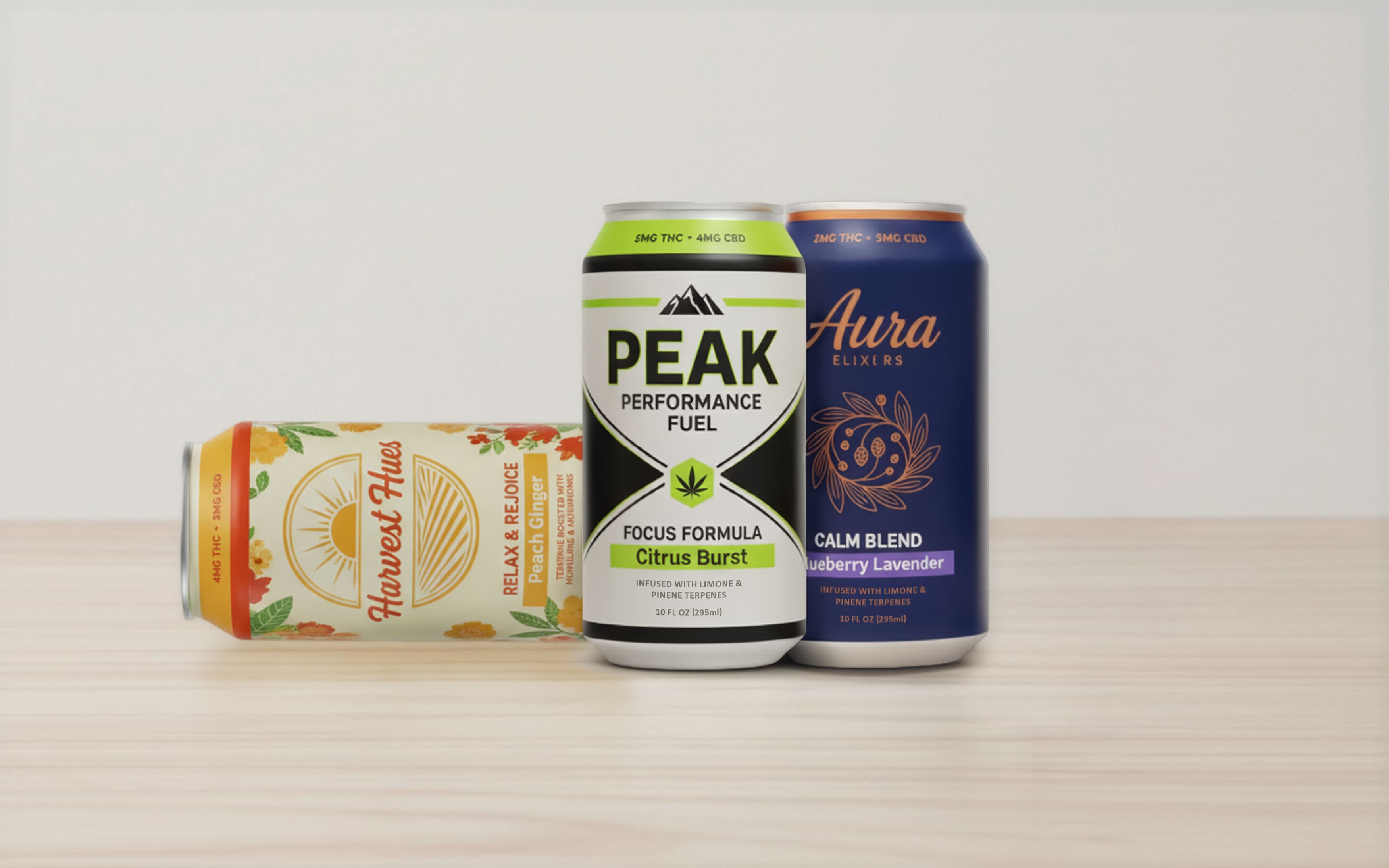 Three beverage cans featuring different flavored elixirs are displayed on a light wooden surface. A cream-colored can lying horizontally is labeled Harvest Hues Peach Ginger. In the center stands a white and lime green can titled PEAK Performance Fuel Citrus Burst. On the right is a dark blue can named Aura Elixirs Blueberry Lavender.