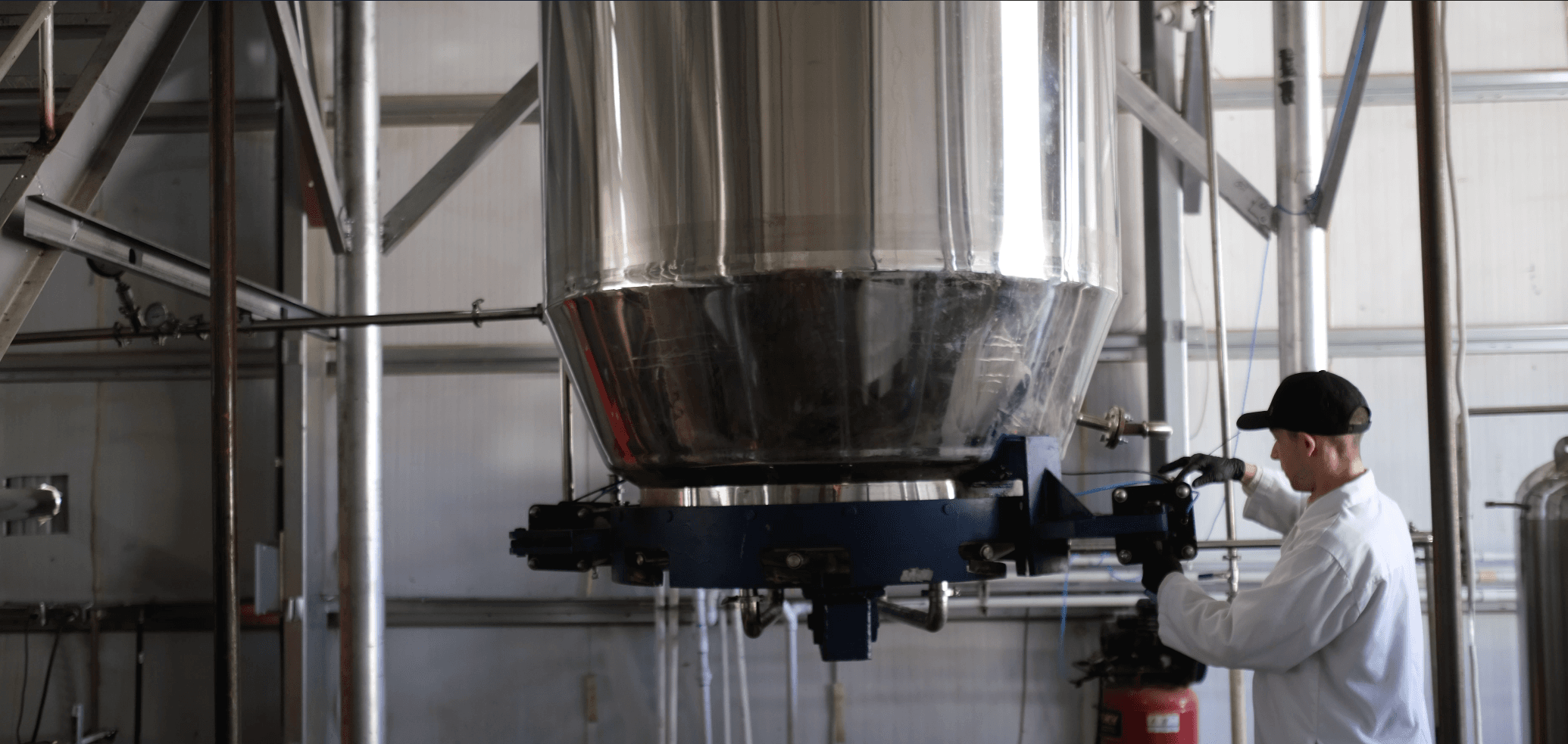 A technician in a white lab coat and black cap operating a large, stainless steel industrial mixing tank in a manufacturing facility.