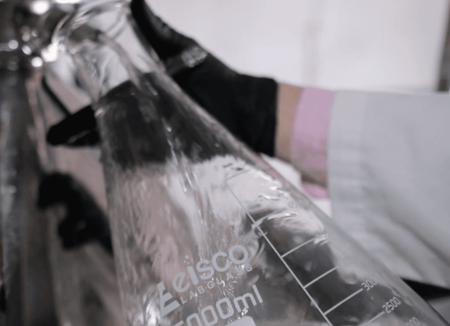 A close-up view of clear liquid being poured into a large Eisco Labglass graduated Erlenmeyer flask held by a person wearing black protective gloves.