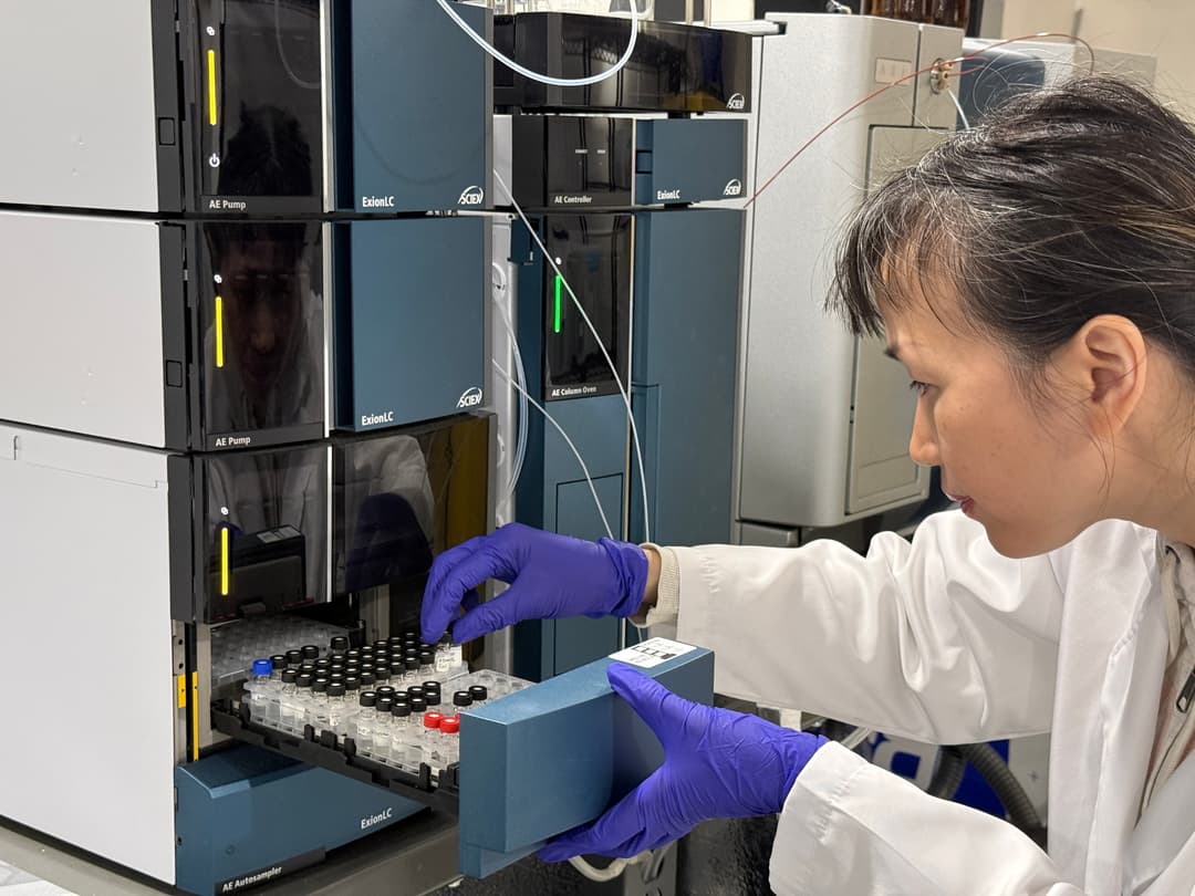 A scientist in a white lab coat and purple nitrile gloves carefully loads small sample vials into an automated liquid chromatography system. She is focused on a tray filled with numerous clear vials, some with black or blue caps, as she slides it into the ExionLC autosampler unit of a large, stacked laboratory instrument