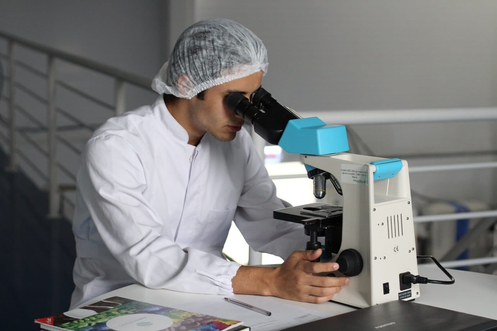 A lab technician wearing a white coat and hairnet uses a high-powered binocular microscope in a clean, professional laboratory setting to inspect product samples.
