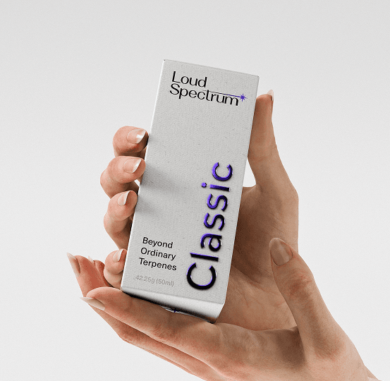 A pair of hands gently cradles a tall, textured light gray box against a plain white background. The packaging features the Loud Spectrum logo at the top and the word Classic printed vertically in a bold, metallic purple font. Text at the bottom reads Beyond Ordinary Terpenes and indicates a 50ml volume.